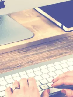 Computer workspace with keyboard, tablet, notebook, water bottle, and iMac at a wooden desk.