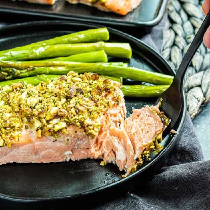 Baked salmon with crushed pistachios, green asparagus, and a black fork on a black plate.