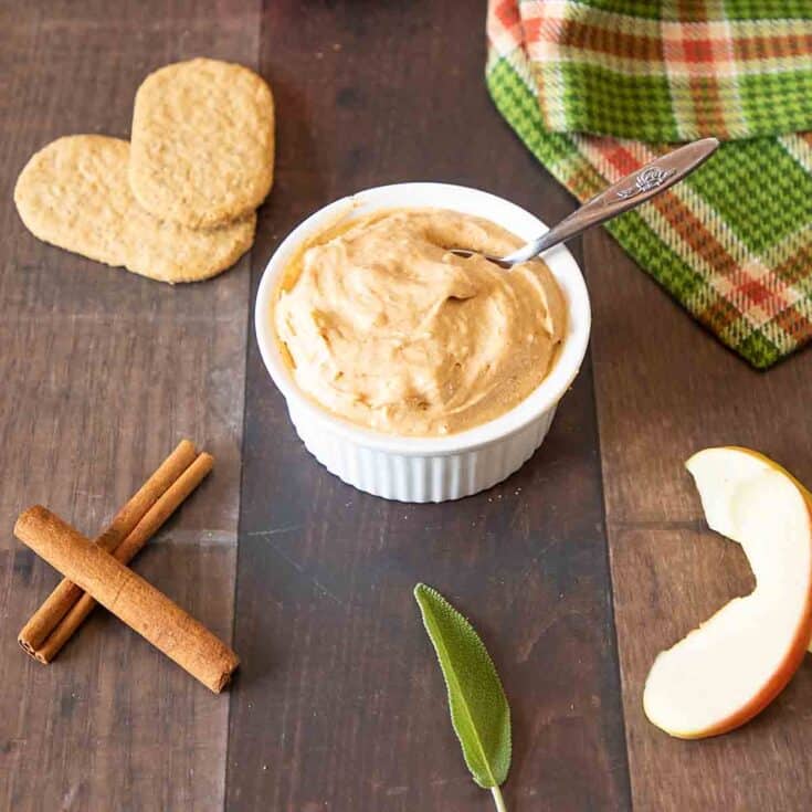 Creamy peanut butter in a white bowl with apple slices and cinnamon sticks on a wooden surface.