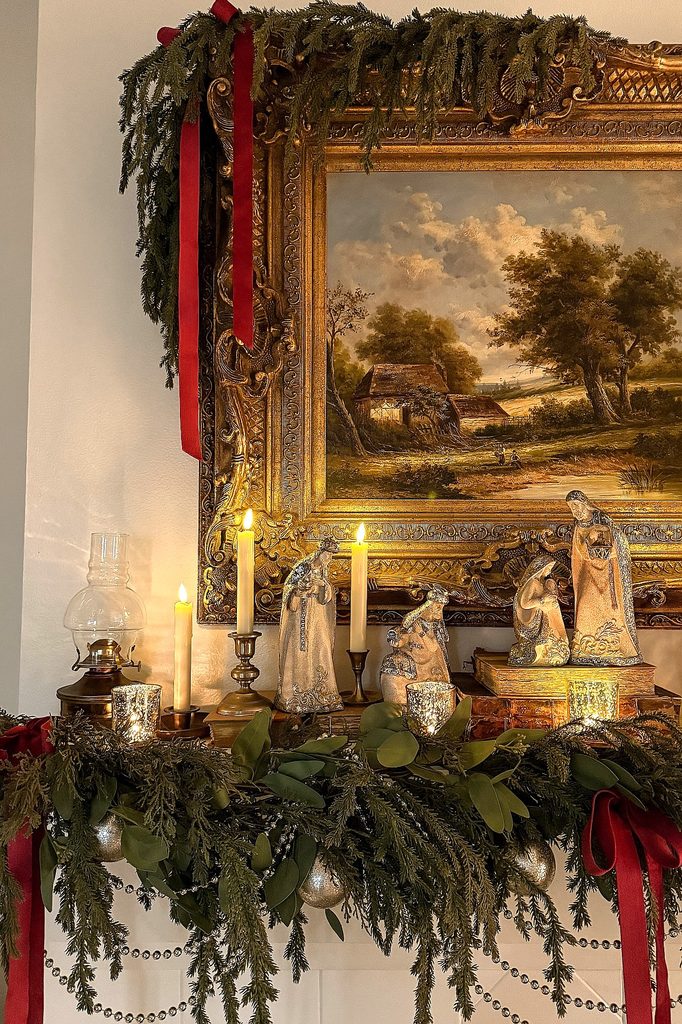 Christmas decorations with candles, greenery, and nativity scene in front of landscape painting.